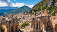 Taormina on Sicily, Italy. Ruins of ancient Greek theater, mount Etna covered with clouds. Taormina old town and mountains in background. 
