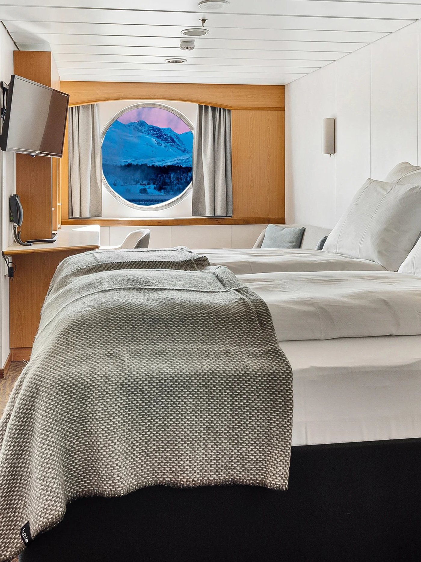 Twin-bedded cabin with porthole window showing coastal views and modern Scandinavian-style furnishings.