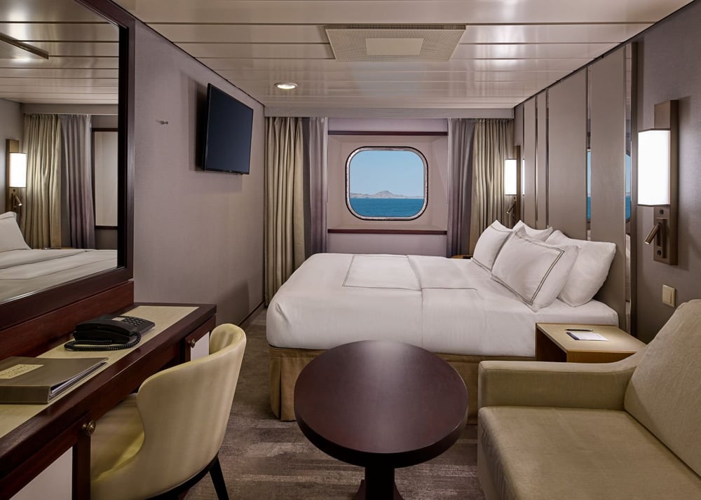 Contemporary cruise ship cabin with queen bed, porthole window, desk, and seating area.