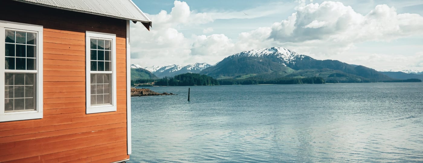 Orange wooden building beside calm water with snow-capped mountain in the background.