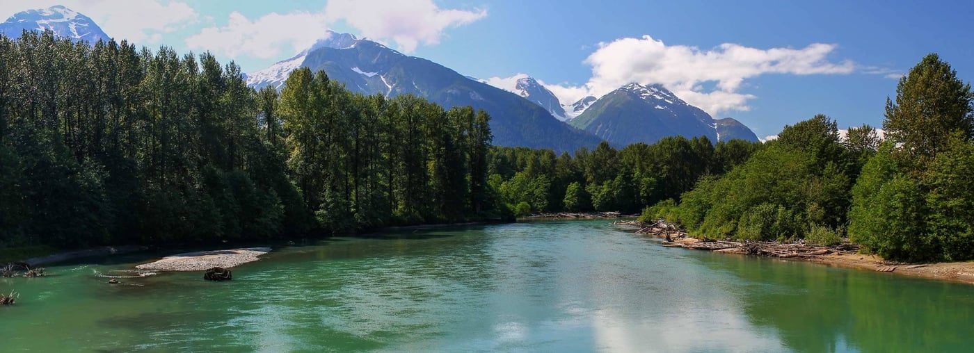 Turquoise river with sandy beach and snow-capped mountains under blue sky.