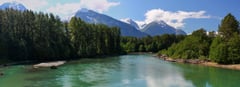 Turquoise river with sandy beach and snow-capped mountains under blue sky.