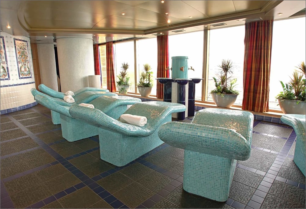 Cruise ship lounge area with turquoise upholstered seating, large windows, and decorative plants in a spacious interior space.