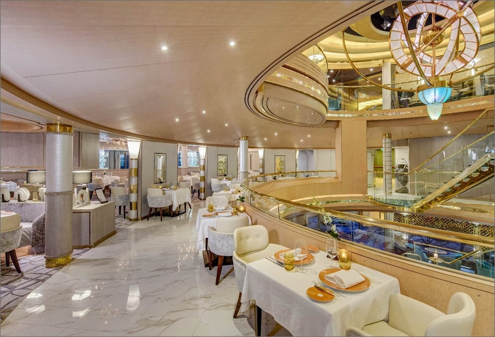 Grand cruise ship dining room with ornate ceiling, chandelier, marble floors, and white-clothed tables.