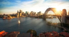 Sydney Harbour at sunset featuring the Opera House, Harbour Bridge, and city skyline with a sailboat.