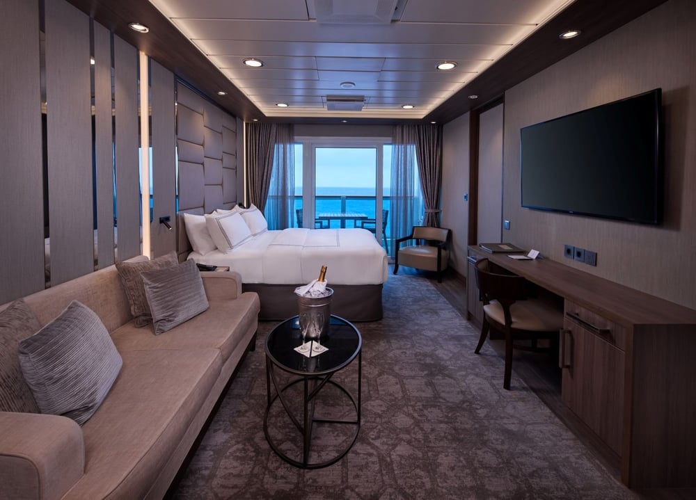Modern cruise ship stateroom with king bed, sofa seating area, balcony access, and wall-mounted television.
