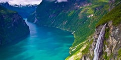 A dramatic Norwegian fjord with turquoise water, steep green mountainsides, and a cascading waterfall.