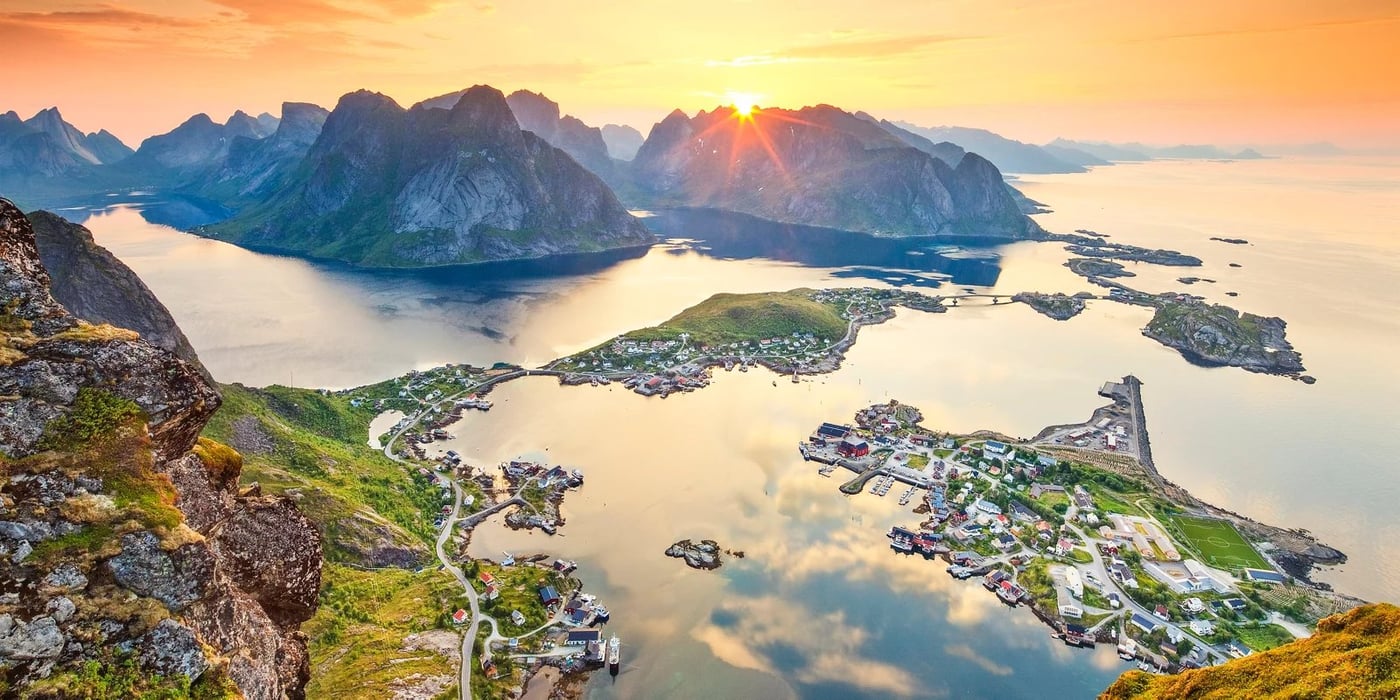 Sunset illuminates dramatic mountain peaks and coastal settlements in Norway's Lofoten Islands from an elevated viewpoint.
