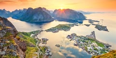 Sunset illuminates dramatic mountain peaks and coastal settlements in Norway's Lofoten Islands from an elevated viewpoint.