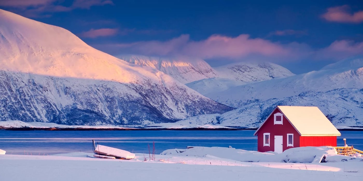 A traditional red Norwegian house sits beside a snow-covered fjord with mountains glowing in pink twilight.