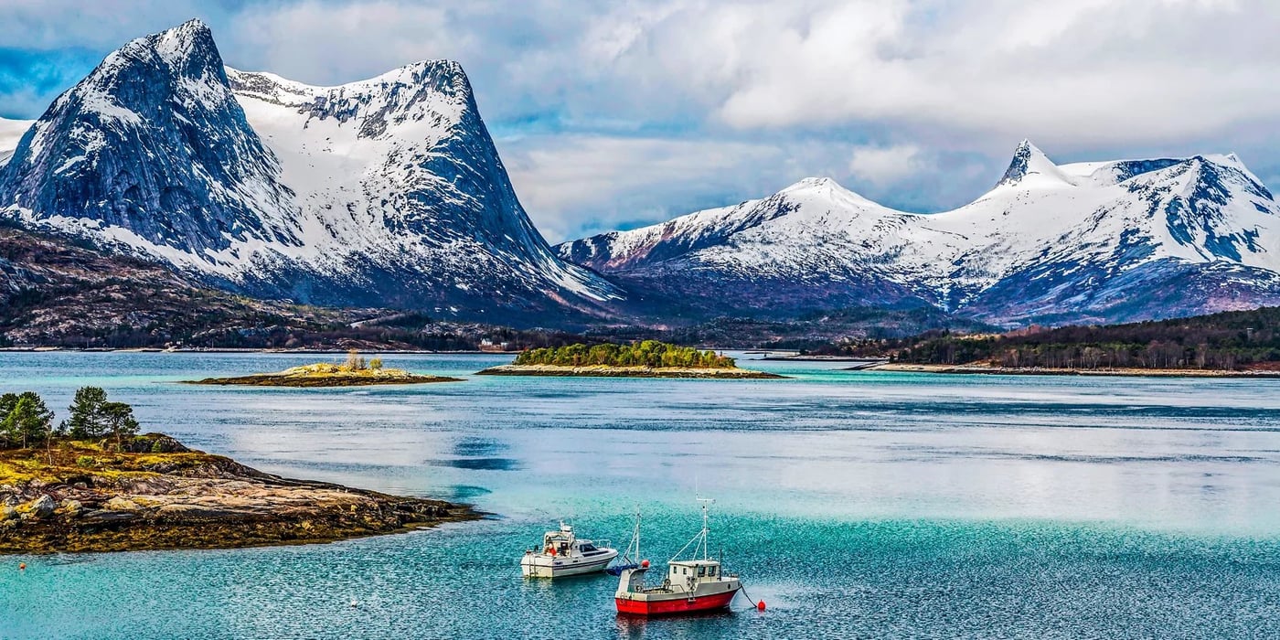 Snow-capped mountains rise above turquoise coastal waters with small boats moored in the foreground in Arctic Norway.