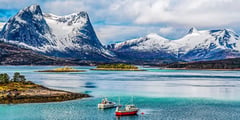 Snow-capped mountains rise above turquoise coastal waters with small boats moored in the foreground in Arctic Norway.