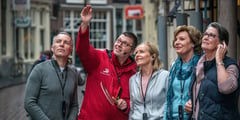 A tour guide in a red jacket points out landmarks to a group of passengers on a European street.