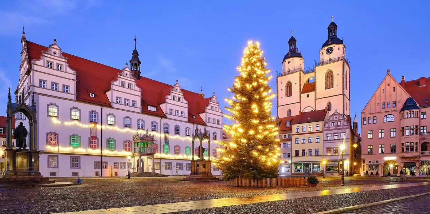 Historic town square at dusk with illuminated Christmas tree and colorful Renaissance buildings reflected on wet cobblestones.