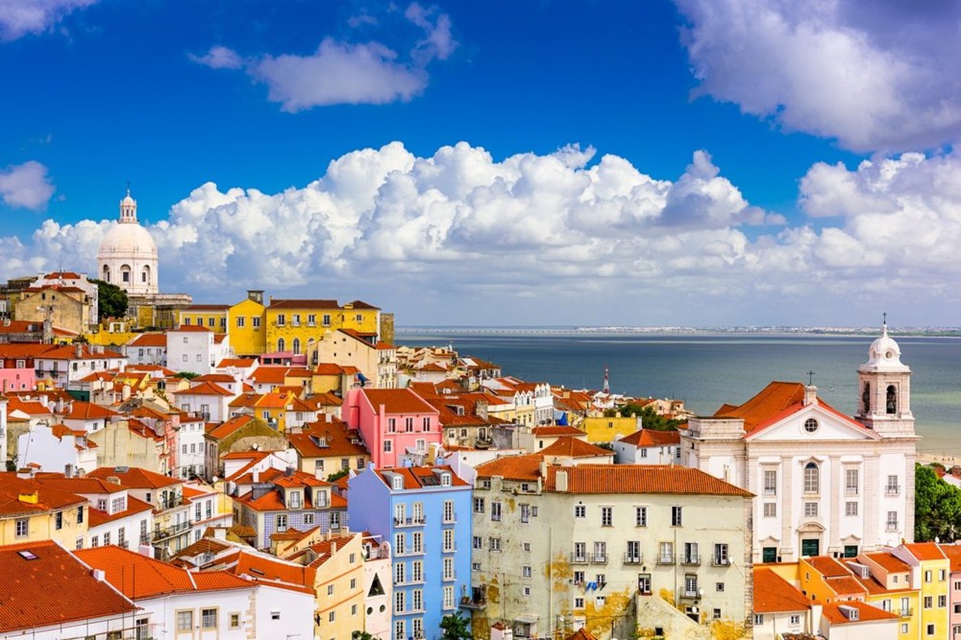 Colorful hillside buildings of Lisbon's Alfama district with terracotta roofs overlooking the Tagus River under blue sky.