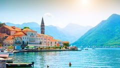 Coastal Mediterranean town with colorful buildings, a church bell tower, and boats moored along the waterfront with mountains in the background.