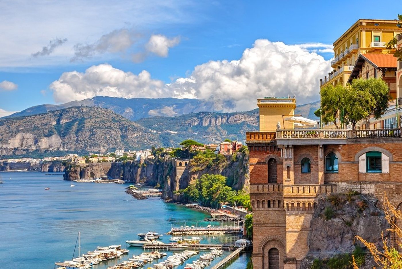 Scenic view of Sorrento coastline with historic buildings perched on cliffs overlooking the Bay of Naples and mountains beyond.
