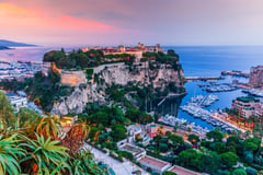 Monaco's Prince's Palace and harbor at sunset with dramatic pink and purple sky, marina, and Mediterranean coastline.