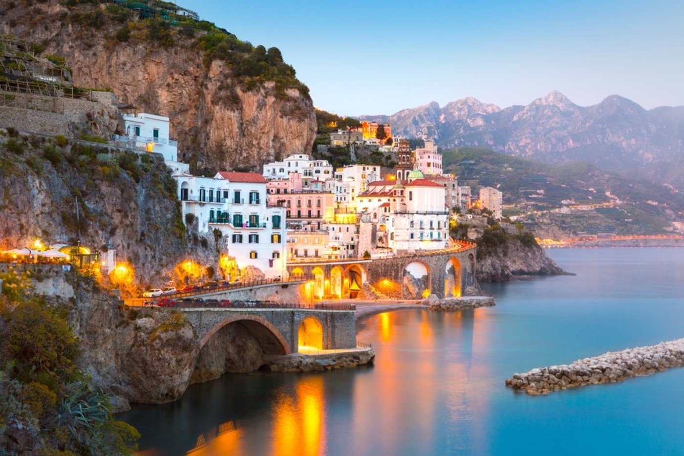 Atrani or Amalfi Coast village at dusk with illuminated buildings cascading down cliffs to the sea and an arched bridge.