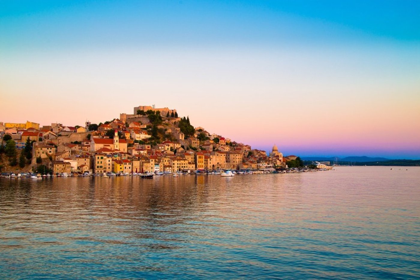 Croatian coastal town built on a hillside overlooking calm waters at sunset with pastel sky.