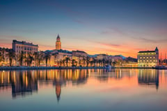 Split waterfront at sunset with illuminated historic buildings and bell tower reflected in the harbor.