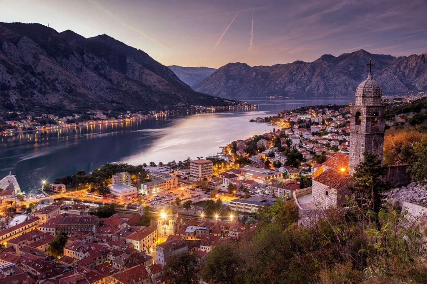 Bay of Kotor at dusk with illuminated waterfront town, church tower, and dramatic mountain backdrop.