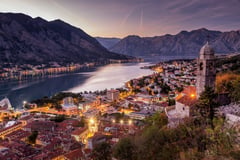 Bay of Kotor at dusk with illuminated waterfront town, church tower, and dramatic mountain backdrop.