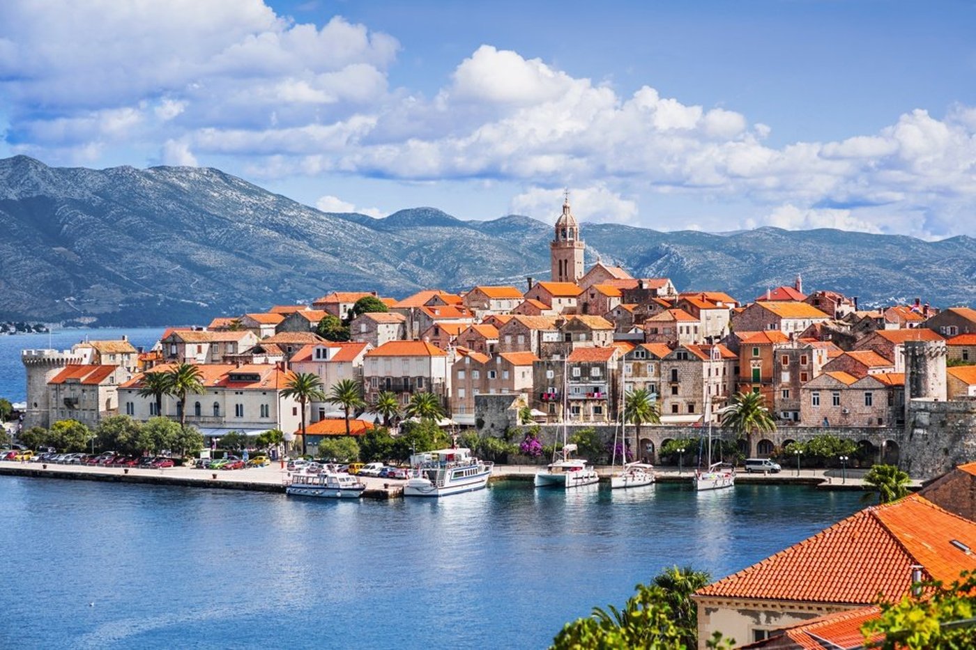 Croatian coastal town with terracotta-roofed buildings and church tower on a peninsula with mountains in the background.