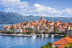 Croatian coastal town with terracotta-roofed buildings and church tower on a peninsula with mountains in the background.