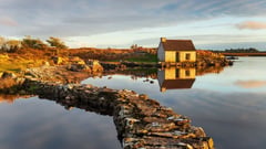 A stone cottage beside a calm lake with rocky shoreline at golden hour in rural Ireland.
