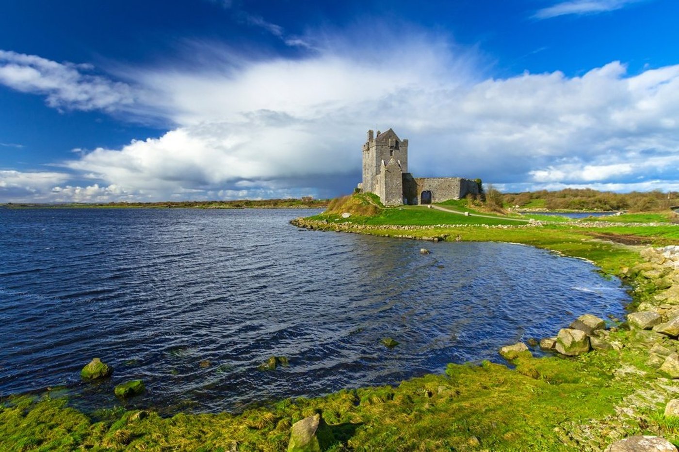 Dunguaire Castle on the shore of Galway Bay with green fields under a dramatic cloudy sky.