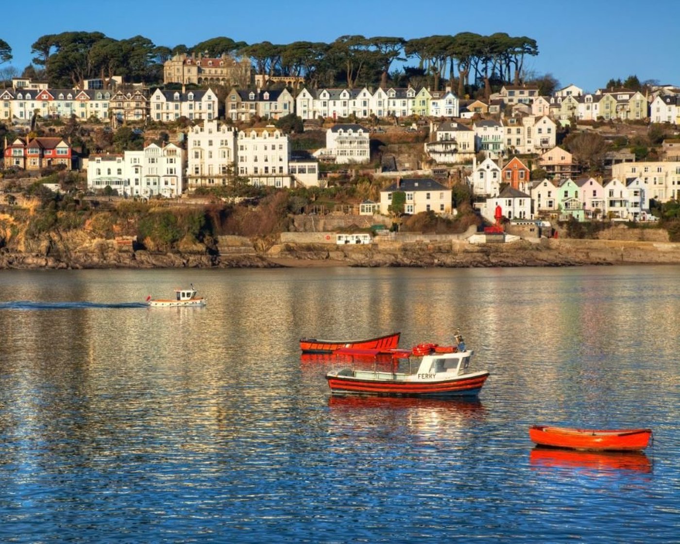 Red fishing boat moored in calm harbor waters with white hillside town and trees in the background.
