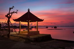 Traditional Balinese pavilion silhouetted against a dramatic pink and purple sunset over calm waters.