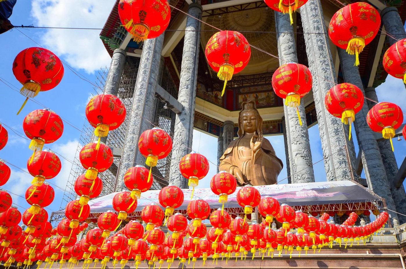 Red Chinese lanterns hanging above a temple entrance with ornate pillars and a Buddha statue.