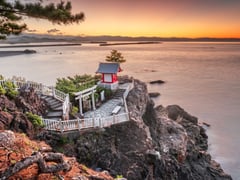 A traditional Japanese shrine with red torii gate perched on rocky coastal cliffs at sunset.
