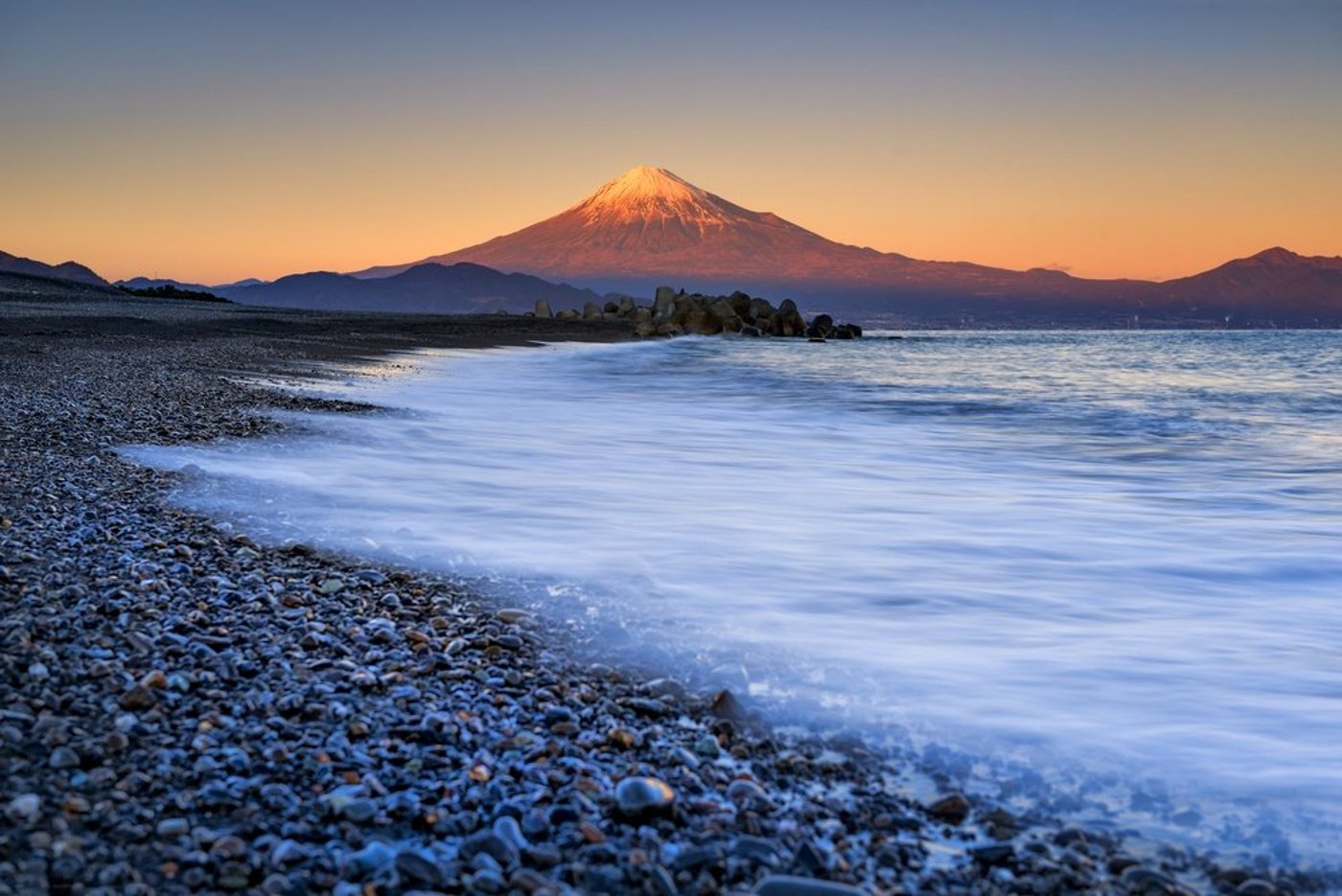 Mount Fuji at sunset viewed from a pebble beach with waves washing ashore.