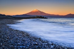 Mount Fuji at sunset viewed from a pebble beach with waves washing ashore.