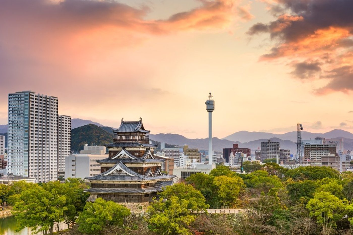 Hiroshima cityscape at dusk showing historic castle amid modern towers with mountains in distance.