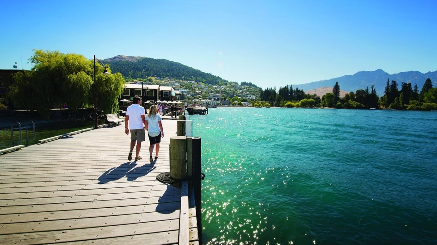 Two people walking on a wooden pier beside a turquoise lake with mountains in the distance.