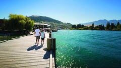Two people walking on a wooden pier beside a turquoise lake with mountains in the distance.