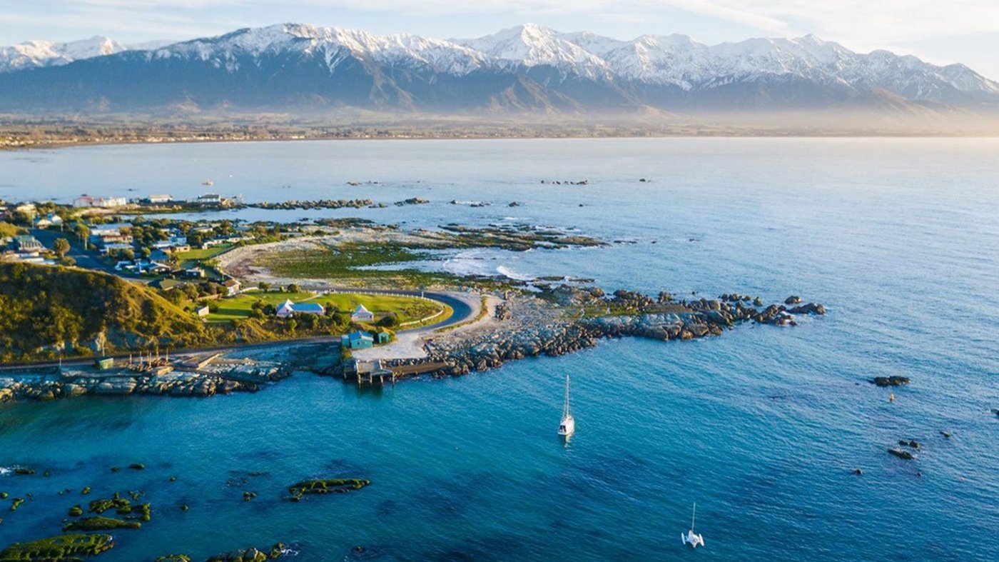 Aerial photograph of a coastal settlement with pier and marina backed by snow-covered mountain range.