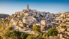 Panoramic view of the ancient hillside town of Matera with its distinctive stone buildings and bell tower under a clear blue sky.