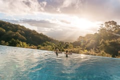 Two people relaxing in an infinity pool overlooking misty hinterland valleys at sunset.