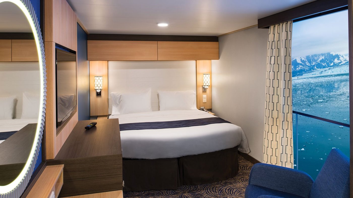 Modern cruise ship cabin interior featuring a queen bed, ocean-view window, and contemporary wood and stone finishes.