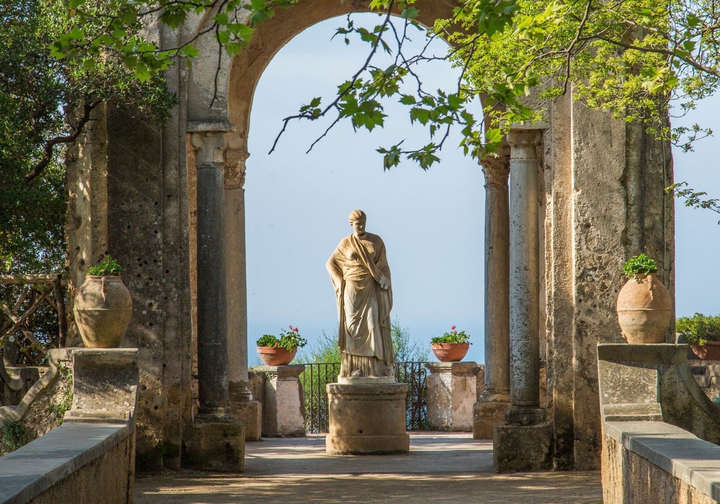 A classical statue framed by ancient columns with coastal views beyond at an Italian garden terrace.