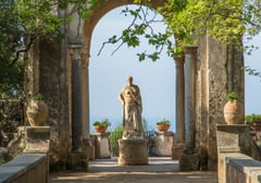 A classical statue framed by ancient columns with coastal views beyond at an Italian garden terrace.